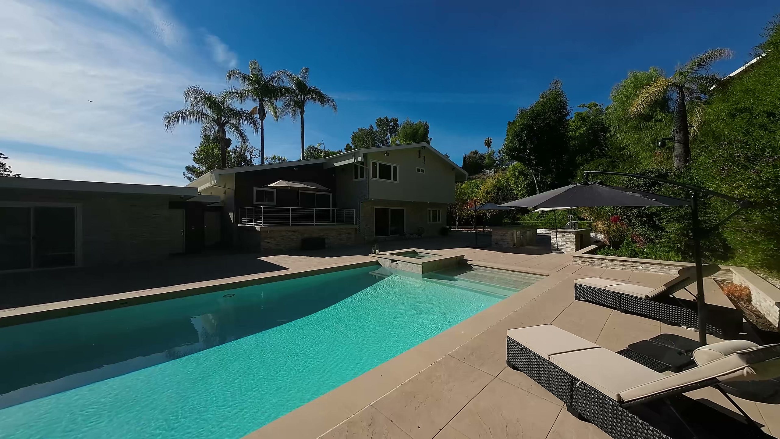 The back of the Winnetka home across the pool, with lounge chairs, umbrellas and tall palms under a blue sky